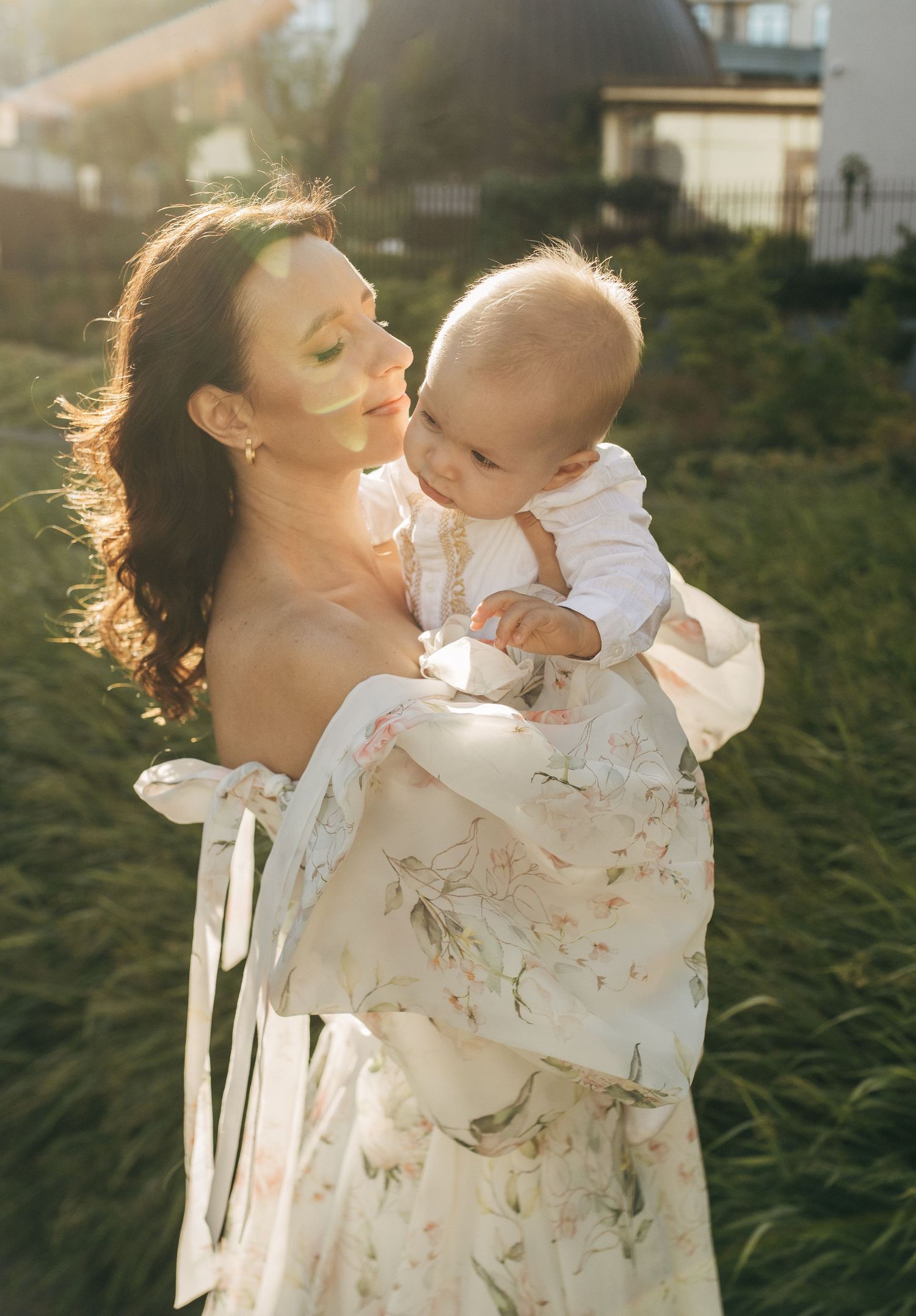 Elegant mother in a floral dress holding her baby in golden sunlight outdoors.