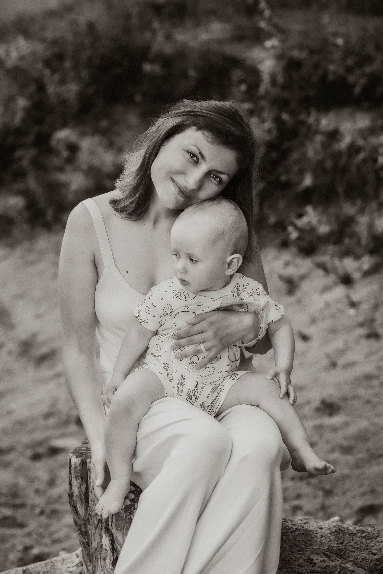 Black and white photo of a smiling mother in white dress holding her baby outdoors on a sunny day.