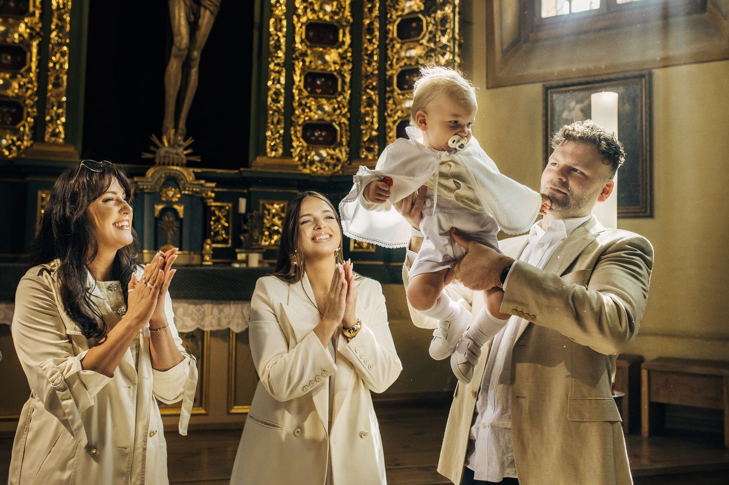 Family celebrating baby baptism in ornate church, parents and godmother in white attire lifting joyful infant.