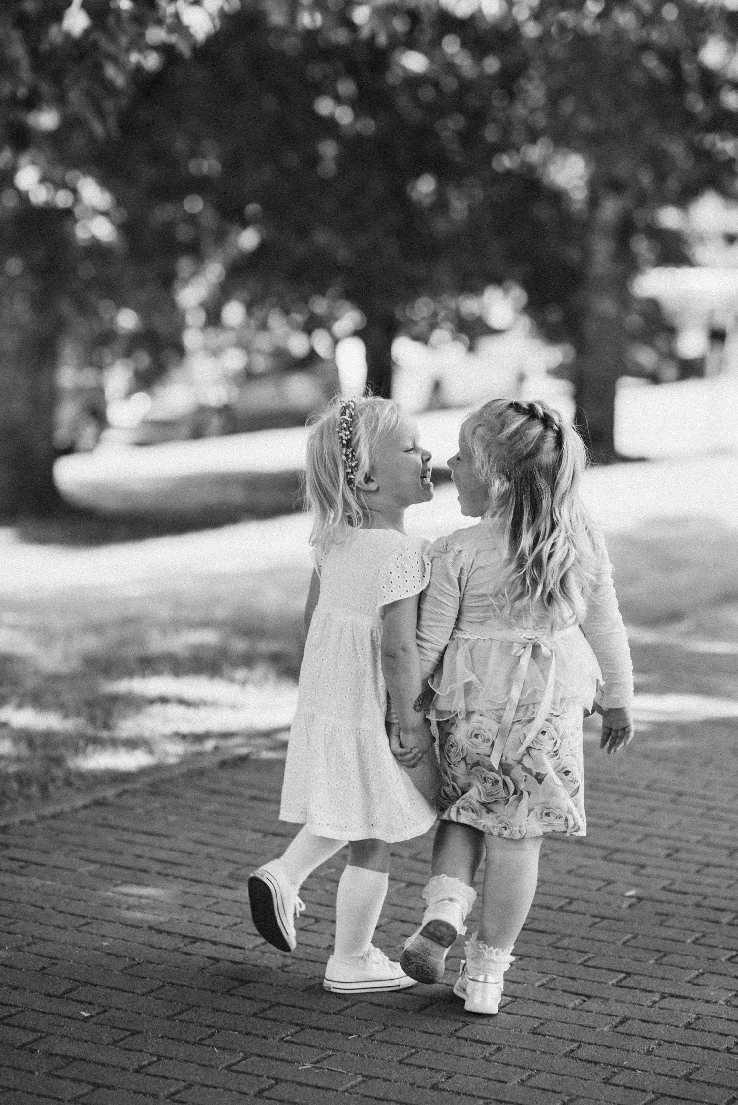 Two little girls in dresses share a sweet kiss on a sunny brick pathway outdoors.