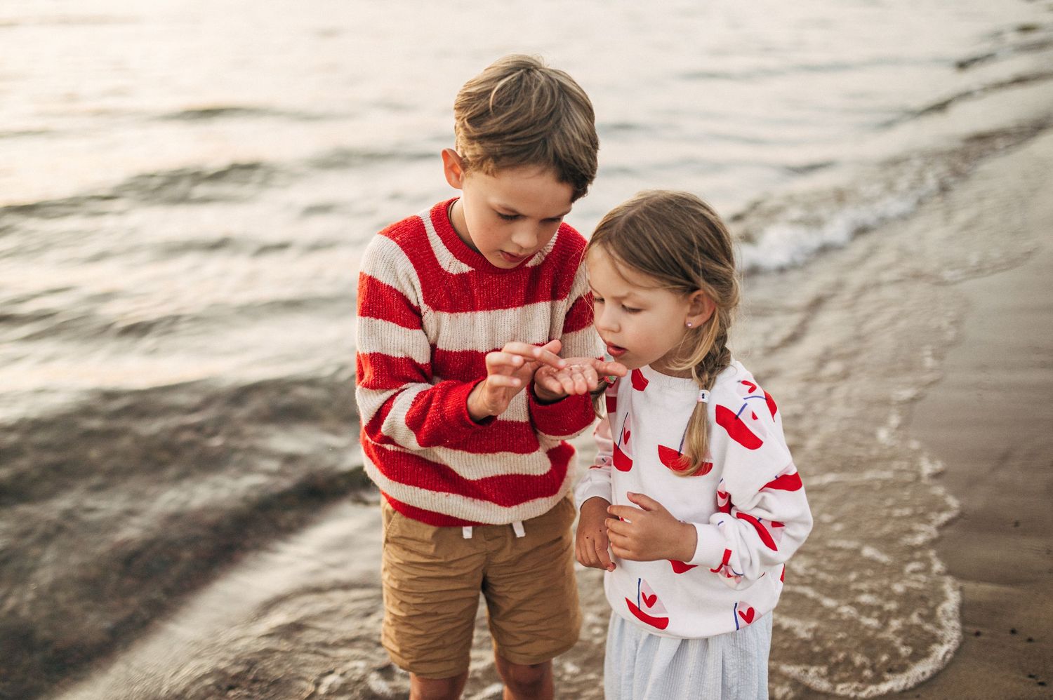 Two children in red and white outfits examining a shell on a sandy beach with ocean waves behind them.