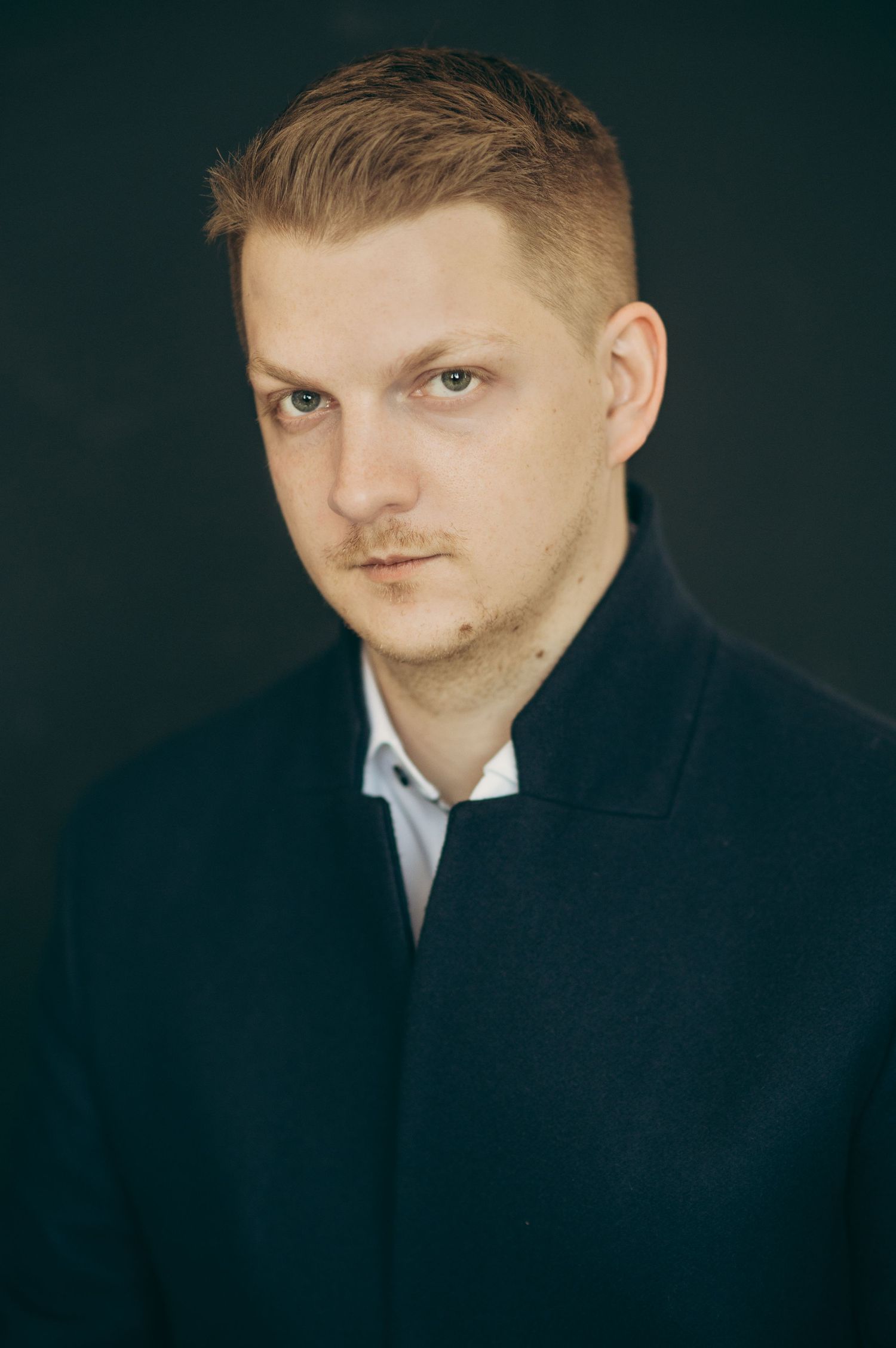 Professional headshot against dark background wearing formal black suit.