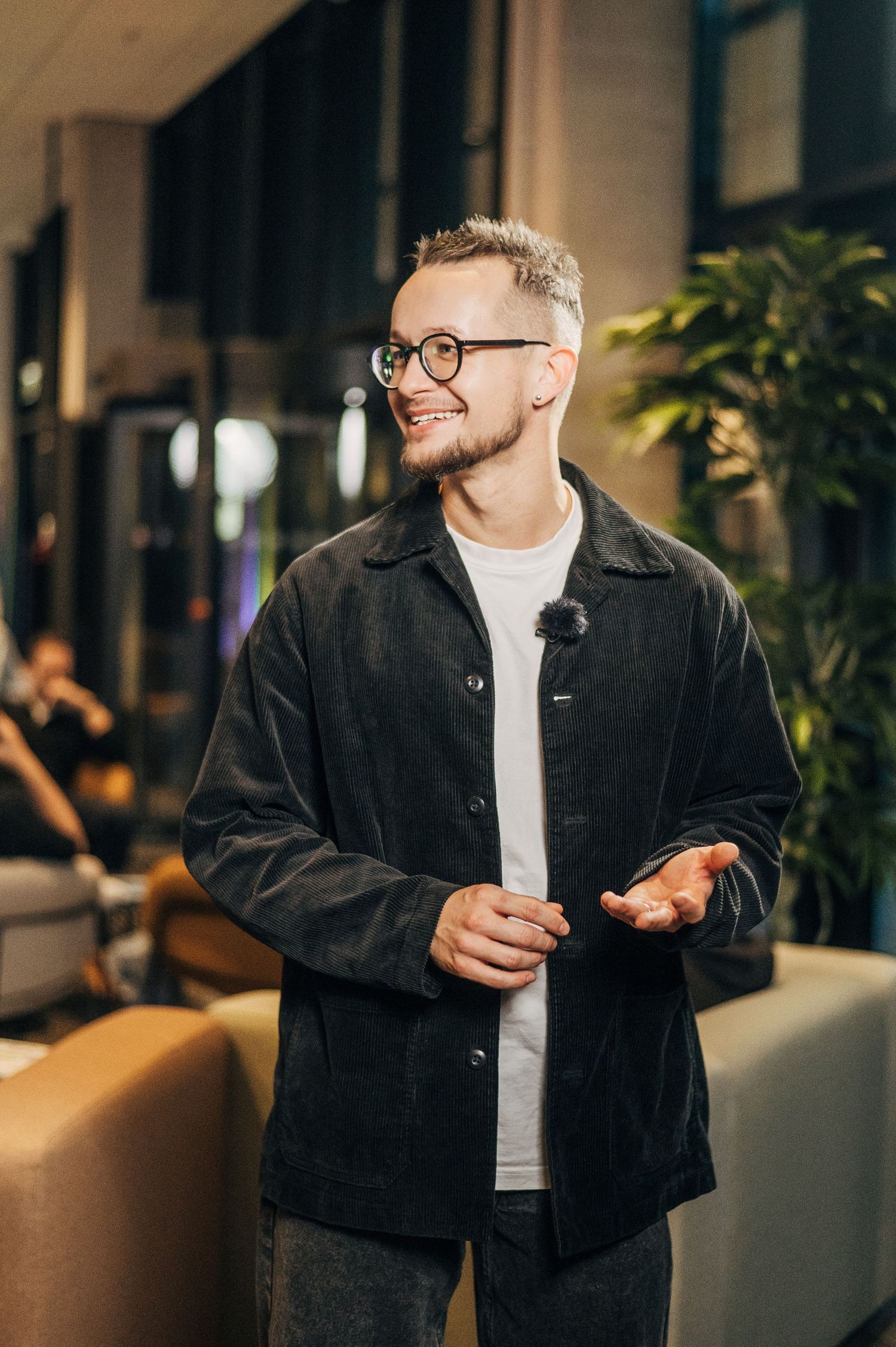Man in black denim jacket and glasses poses in modern interior with soft lighting and plants.