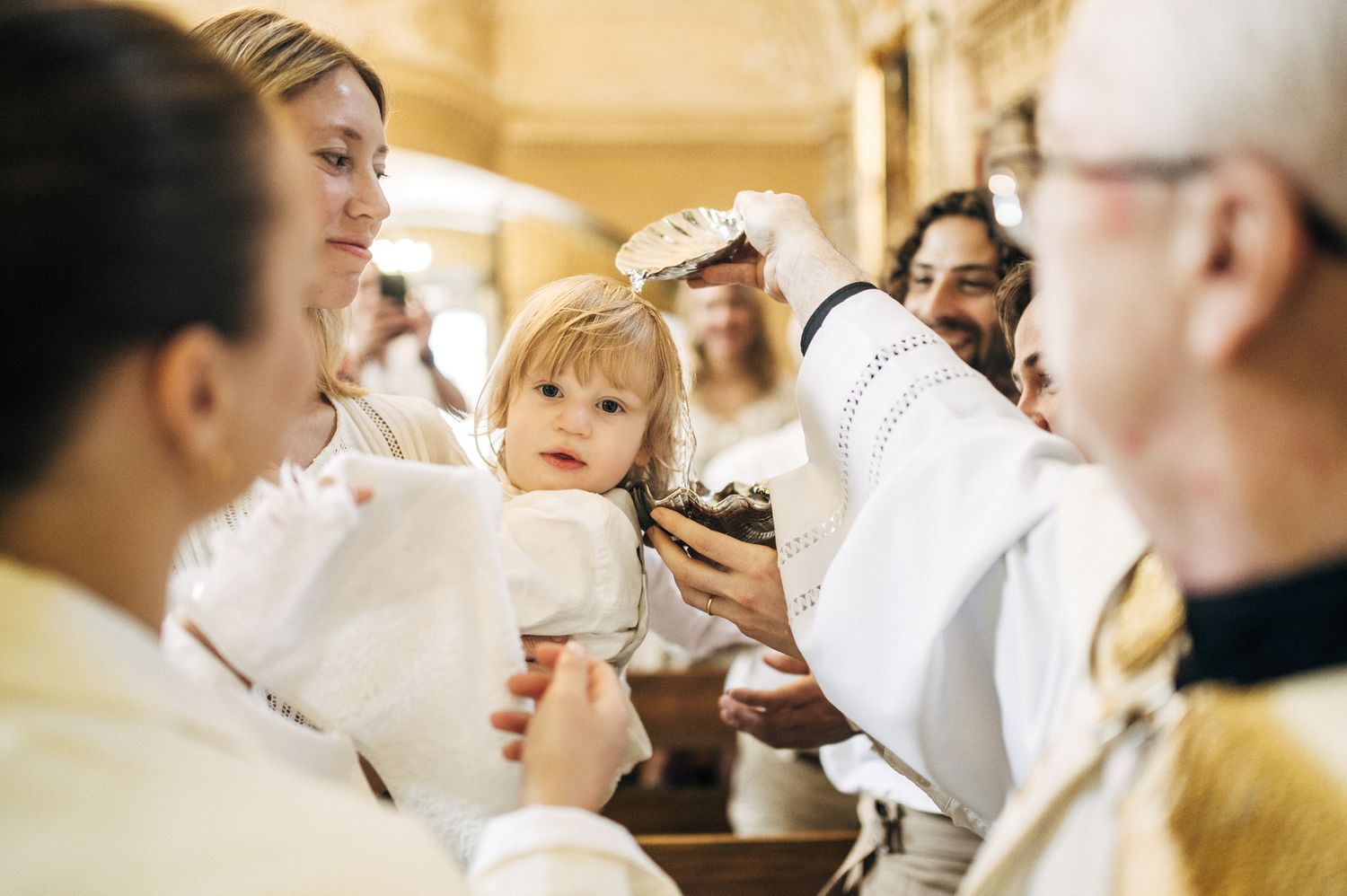 Priest performing baptism on a young child held by a woman in a church.