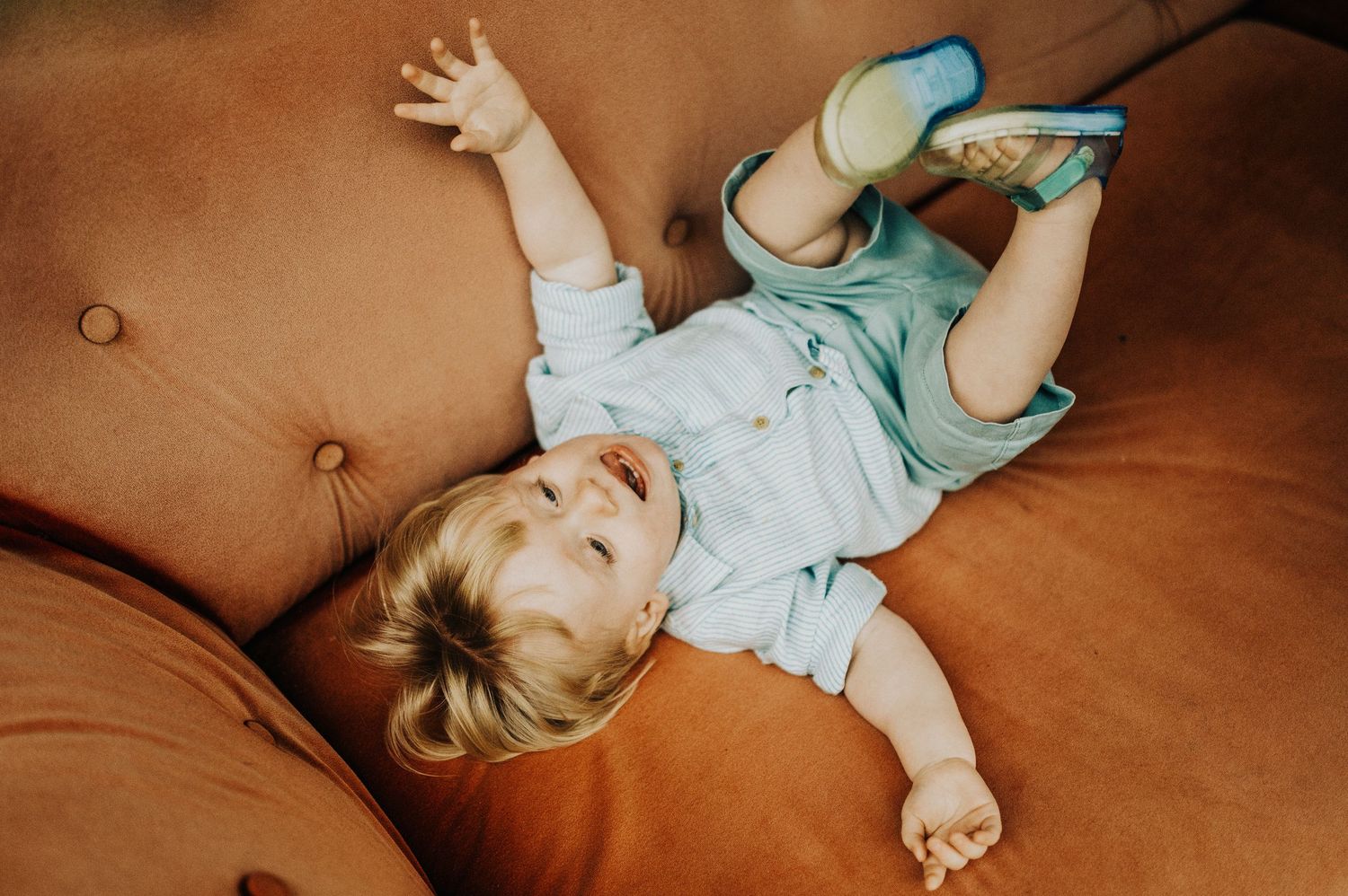 Happy baby lying on a brown tufted sofa wearing a light blue outfit and colorful shoes.