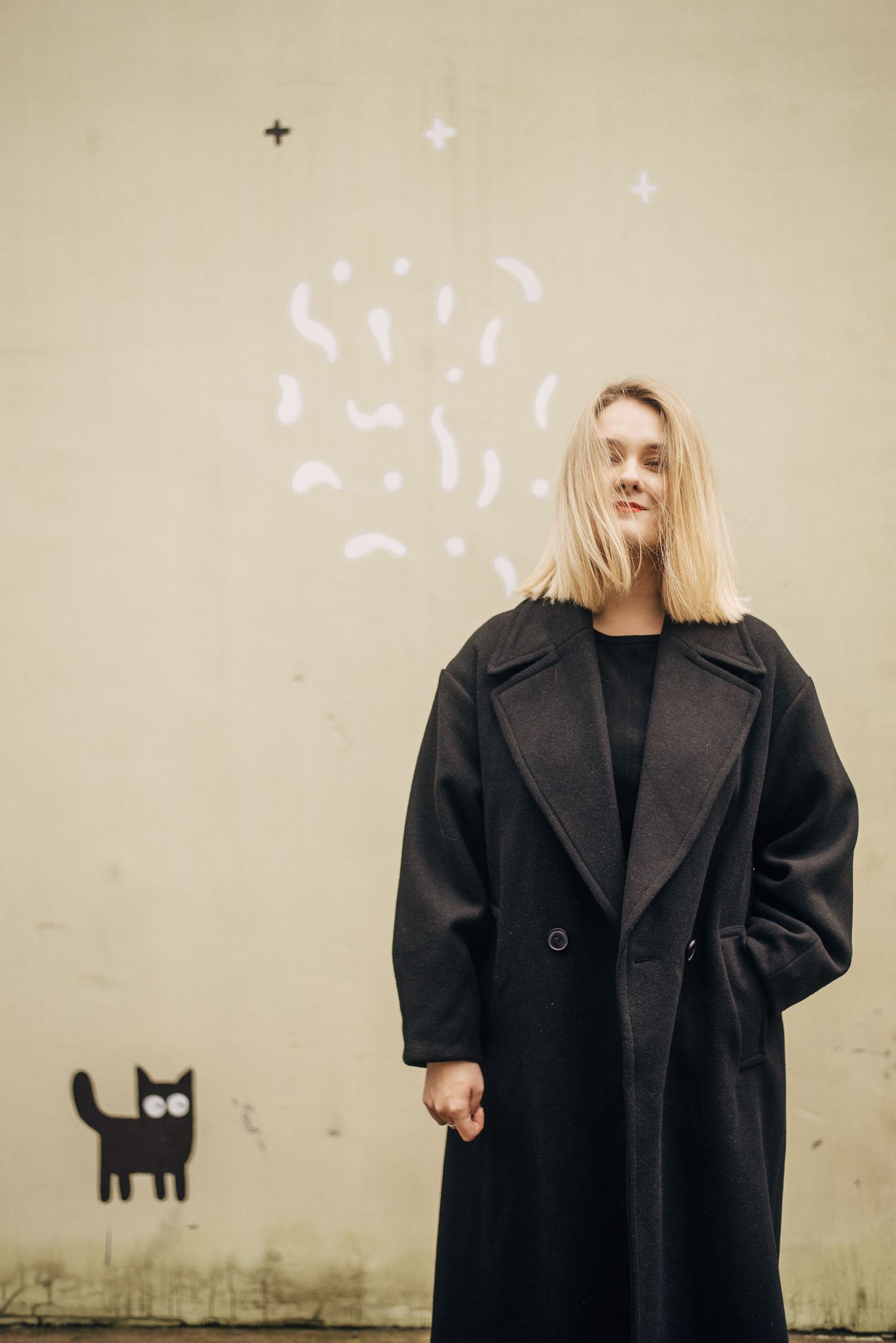Person in black oversized coat against wall with cat graffiti and floating light effects.