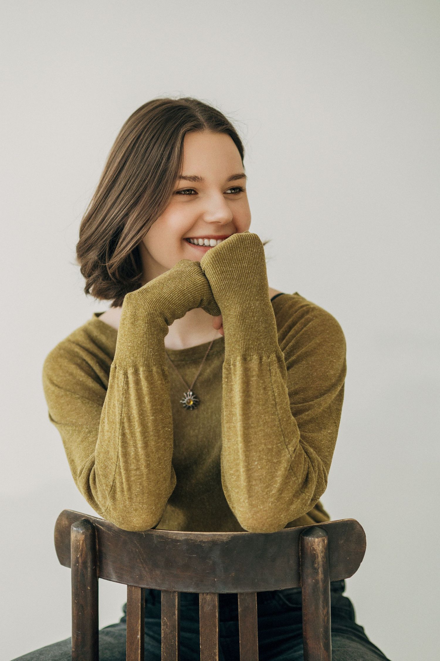 Person in olive sweater shows playful expressions while posing on a wooden chair.