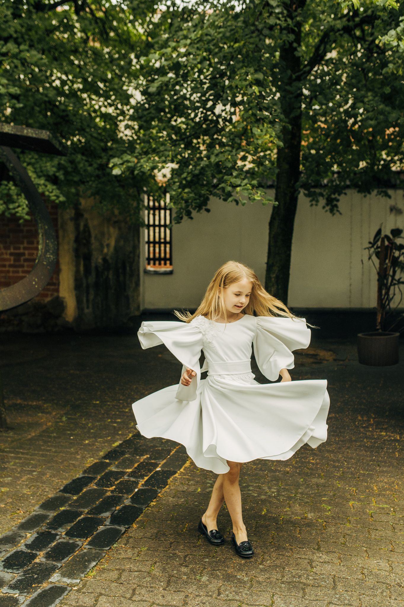 Young blonde girl twirling in white puff-sleeve dress in outdoor courtyard.