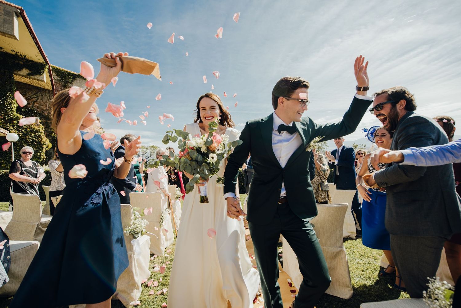 Wedding party celebration with flower petals being tossed against a bright blue sky.
