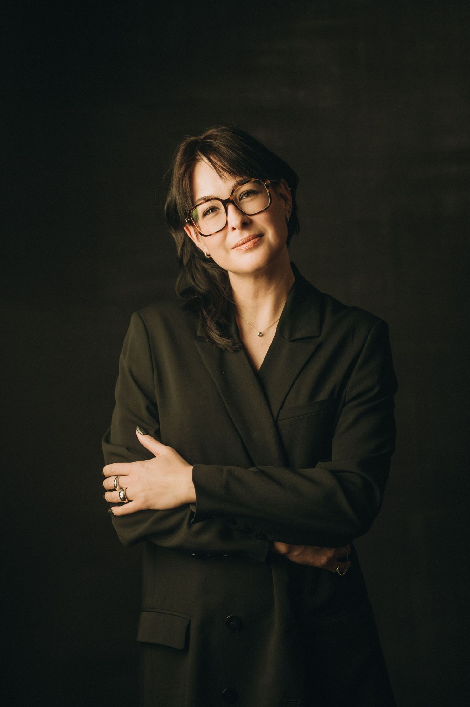 Warm portrait of person in dark blazer and glasses smiling against moody background.