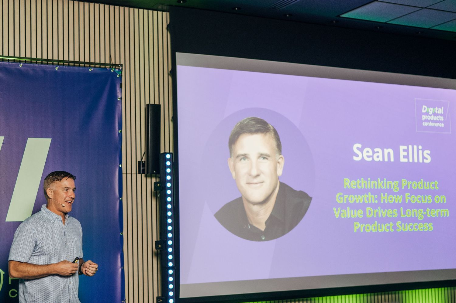 Speaker presenting on stage at a professional conference with purple lighting and presentation slides visible.
