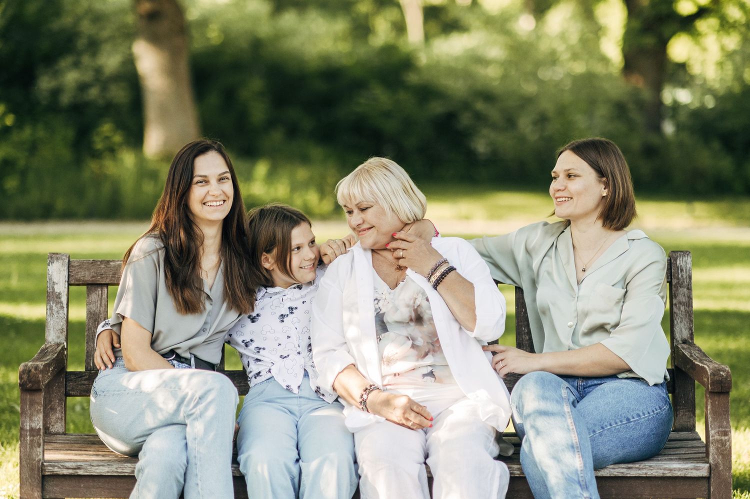 Three people sit together on a wooden bench in a sunlit garden setting.