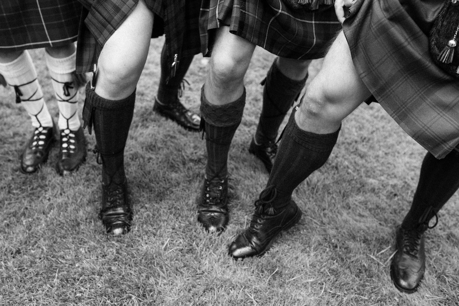 Black and white photo showing legs in traditional Scottish kilts and dress socks at a formal gathering.