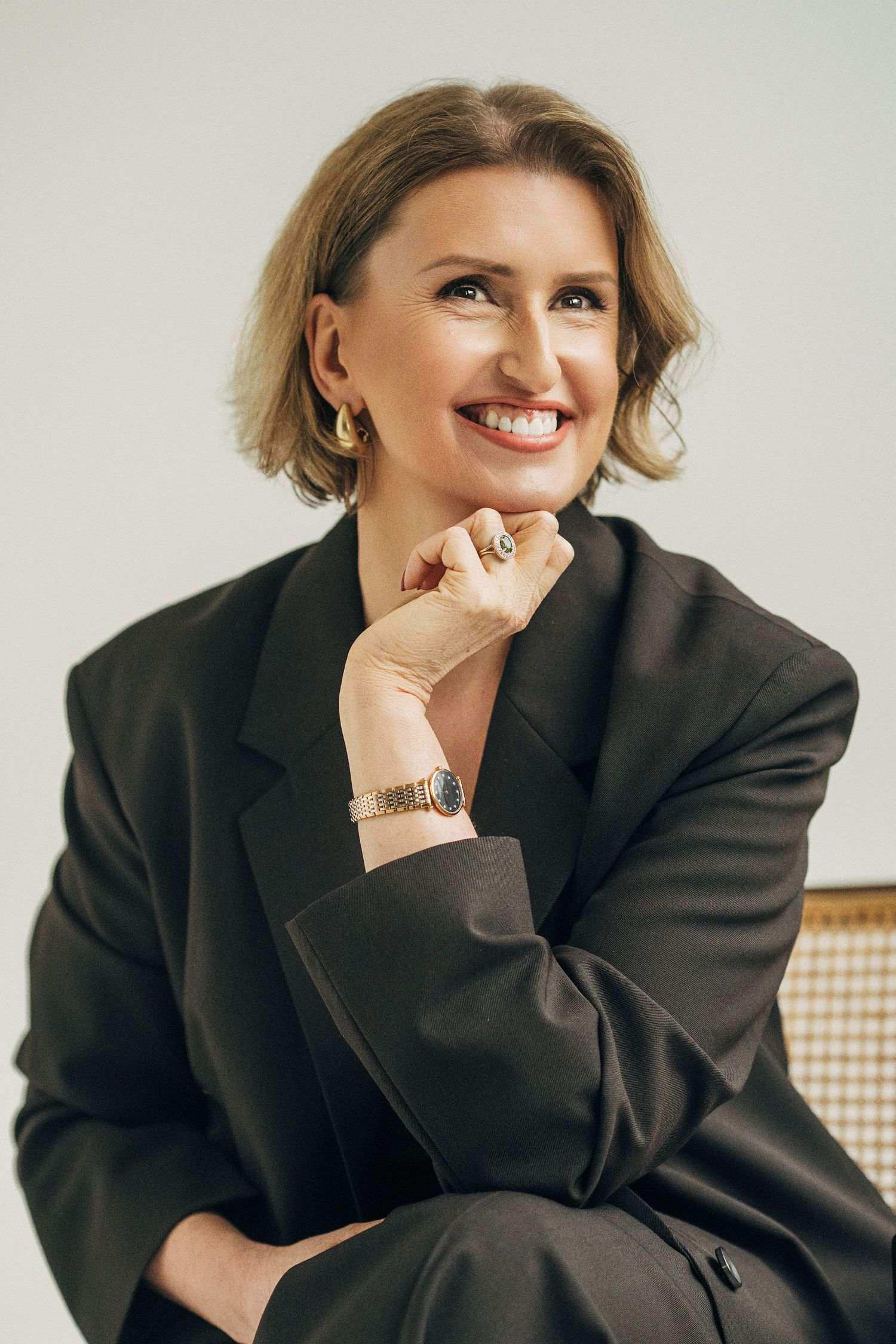 Professional portrait of someone in a black blazer smiling warmly while wearing gold earrings and a watch.