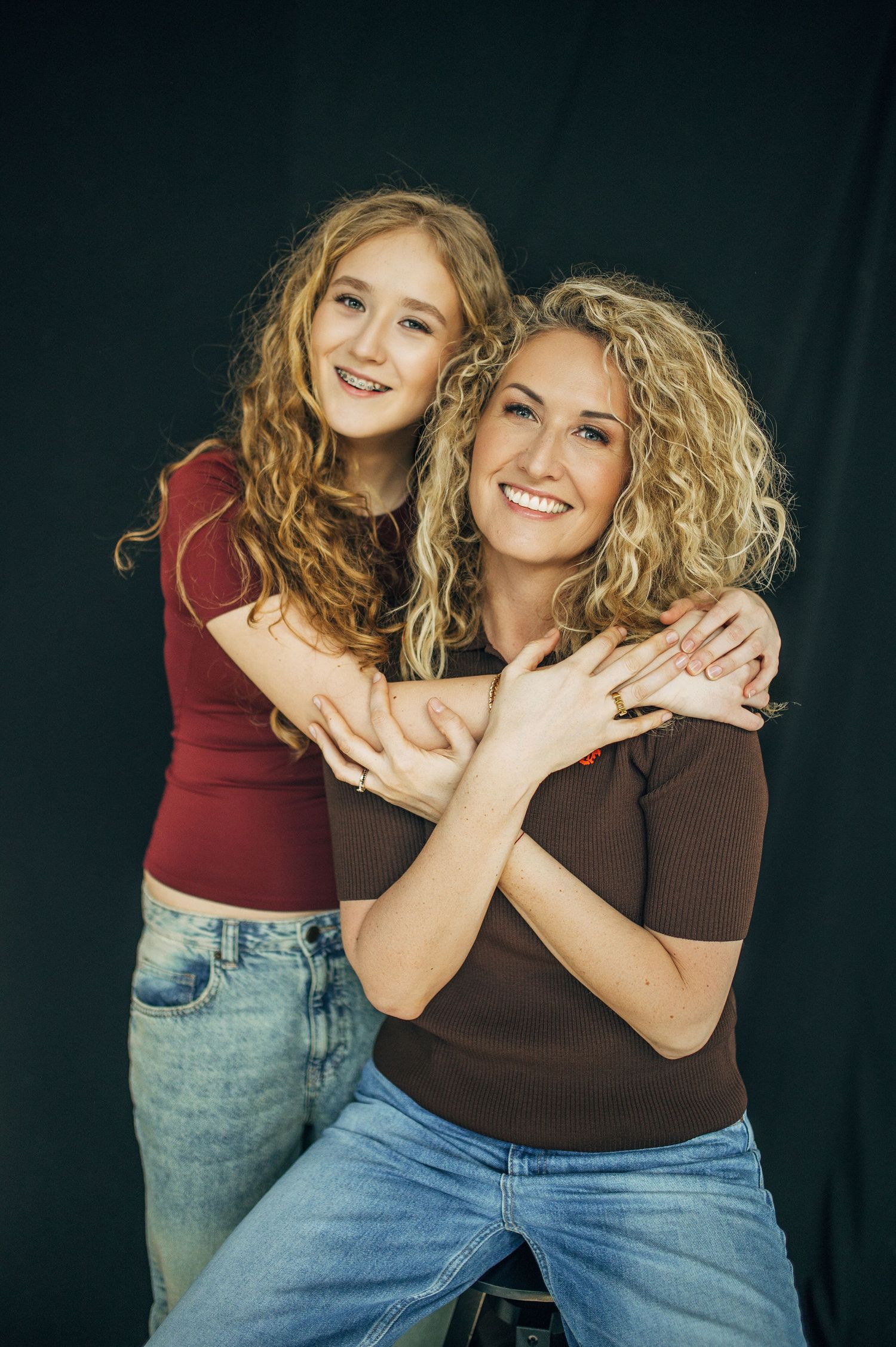 Two curly-haired women smiling and hugging in a studio, wearing casual tops and jeans against a dark background.