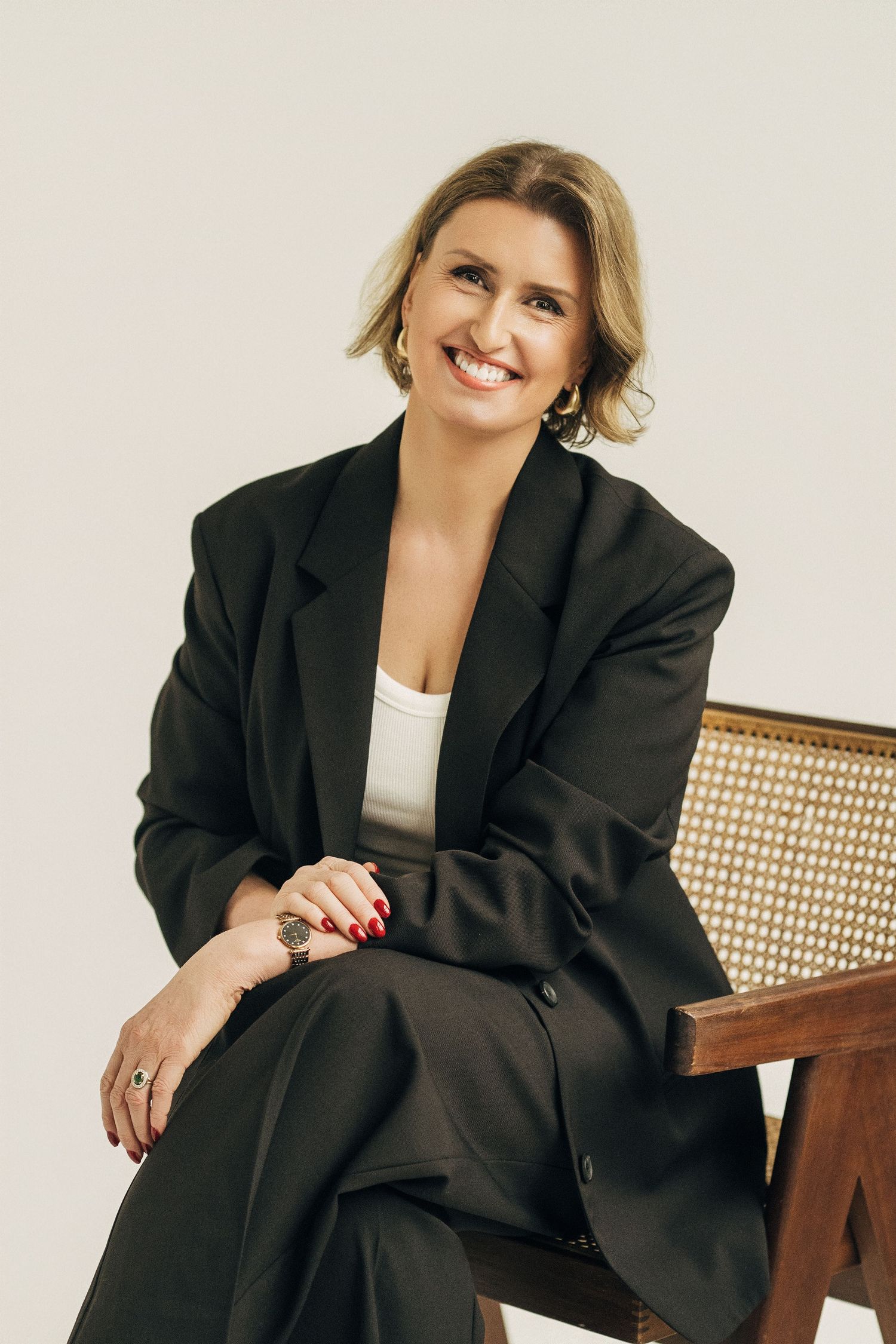 Professional portrait of someone in a black blazer sitting on a wooden chair with cane backing against white backdrop.