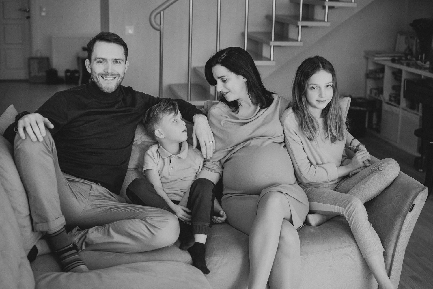 A family of four relaxes together on a gray couch in a black and white photo.