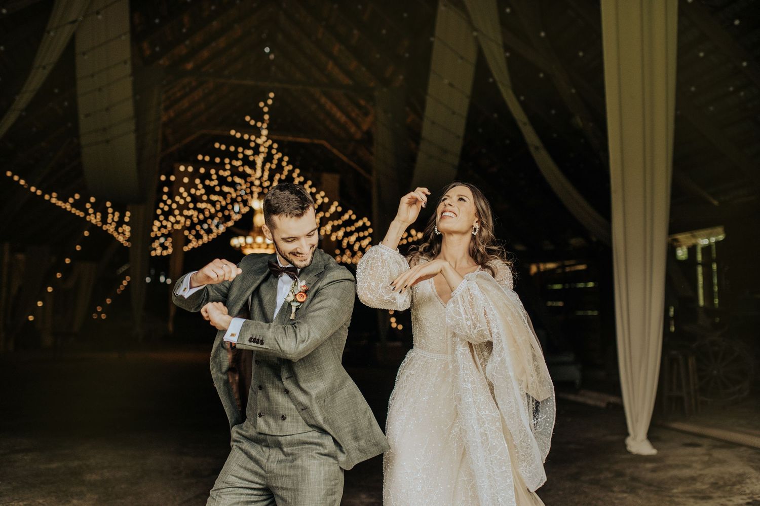 A couple dancing joyfully at their wedding reception under string lights in a rustic venue.