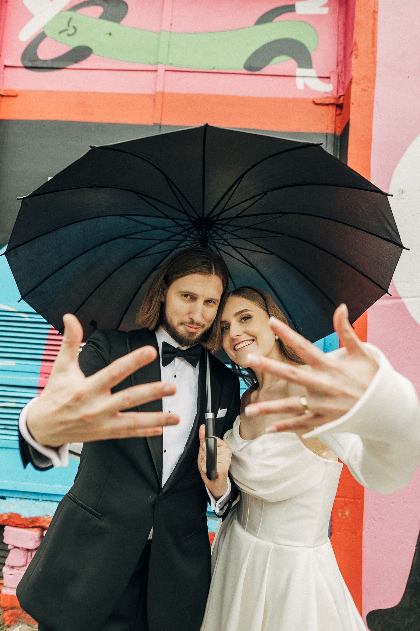 A couple in formal attire sharing a romantic moment under a black umbrella against a pink and red wall background.