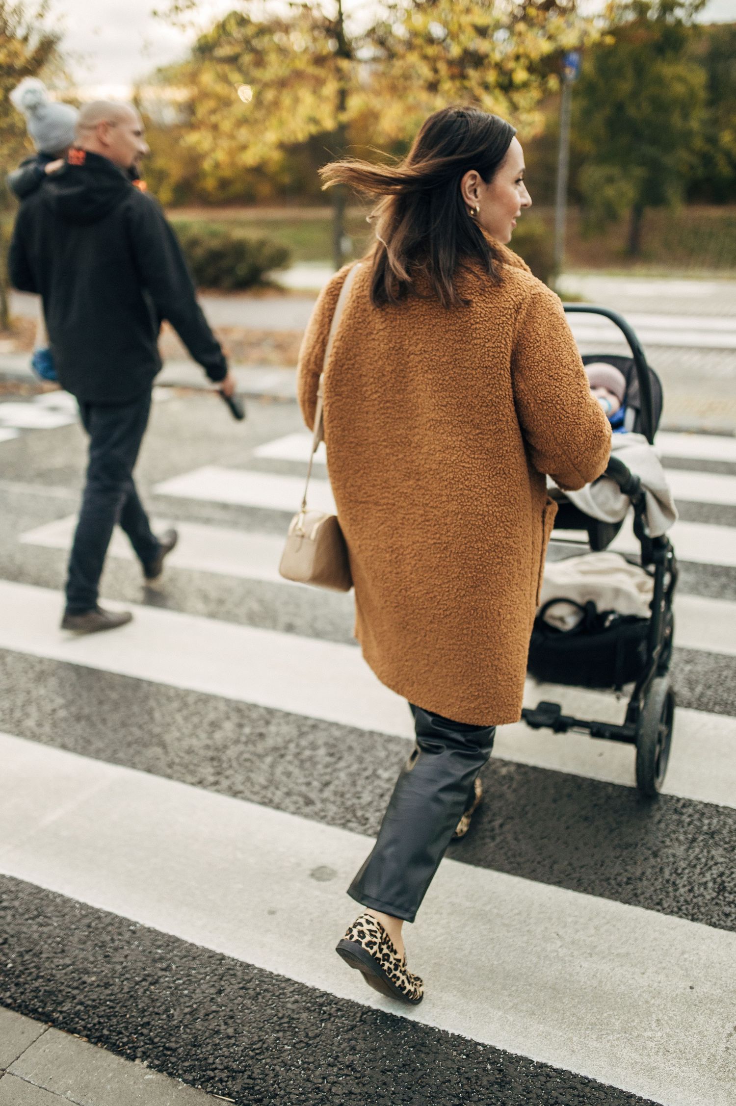 A person wearing a brown sweater walks along a crosswalk path on an autumn day.