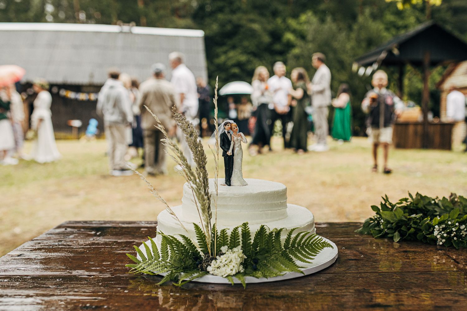 Outdoor wedding reception setup with rustic wooden tables, greenery decor, and guests mingling in a sunny garden setting.