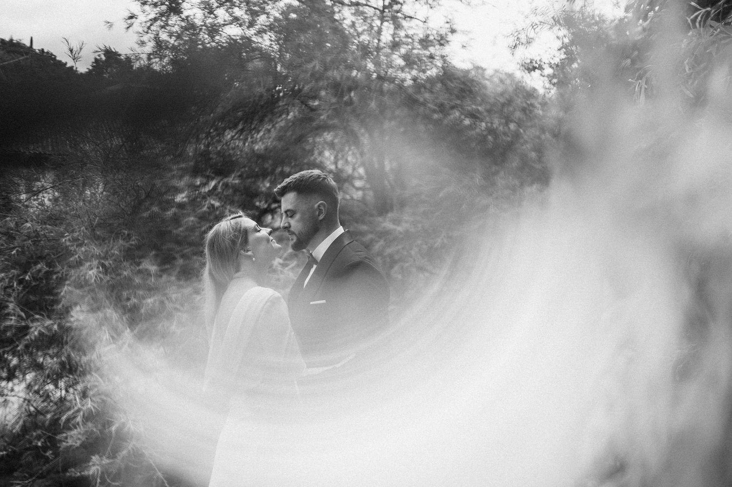 Black and white wedding photo of bride and groom sharing a romantic moment surrounded by lush greenery.