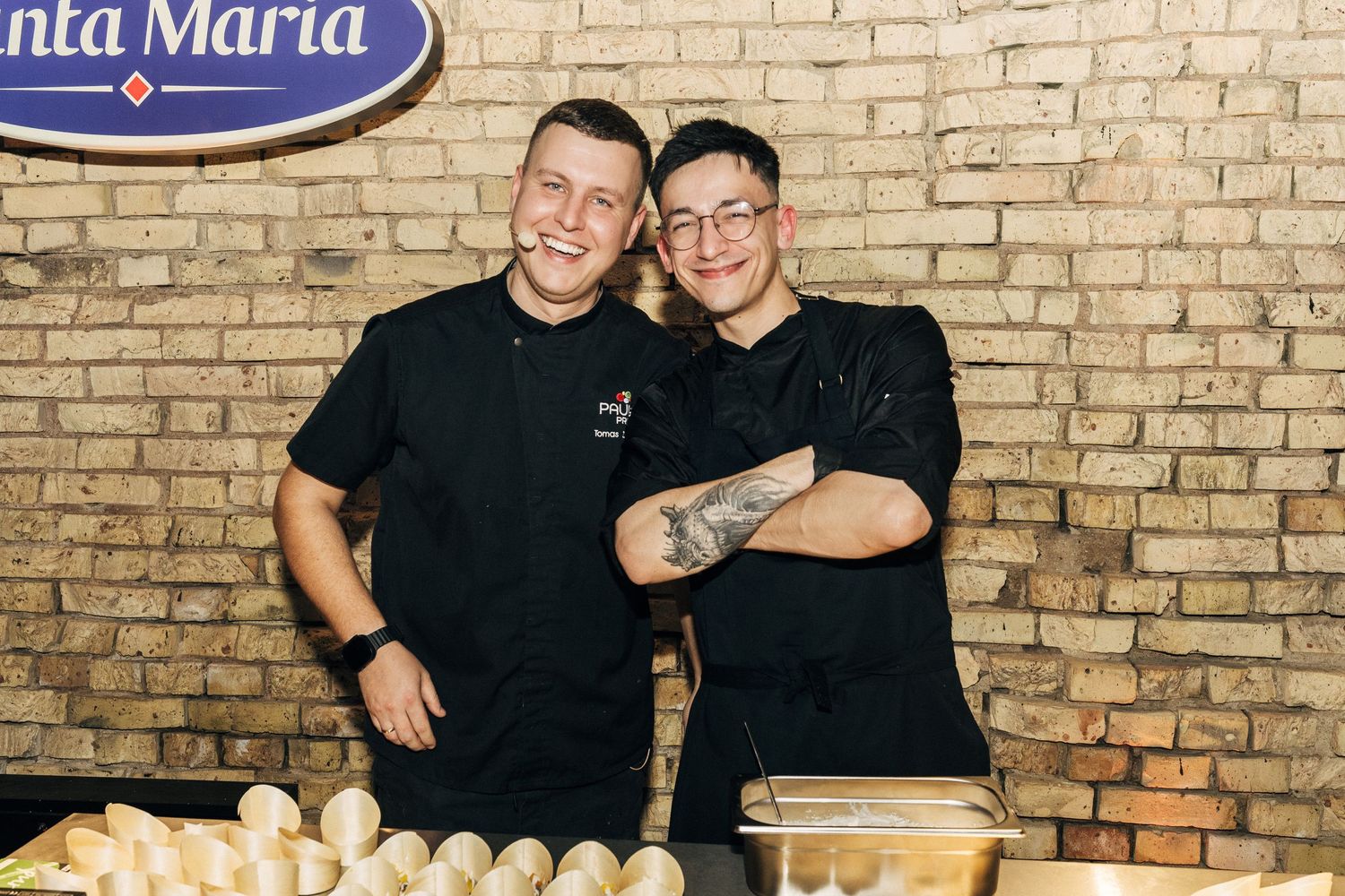 Two people stand together in front of a brick wall in a restaurant or bar setting.