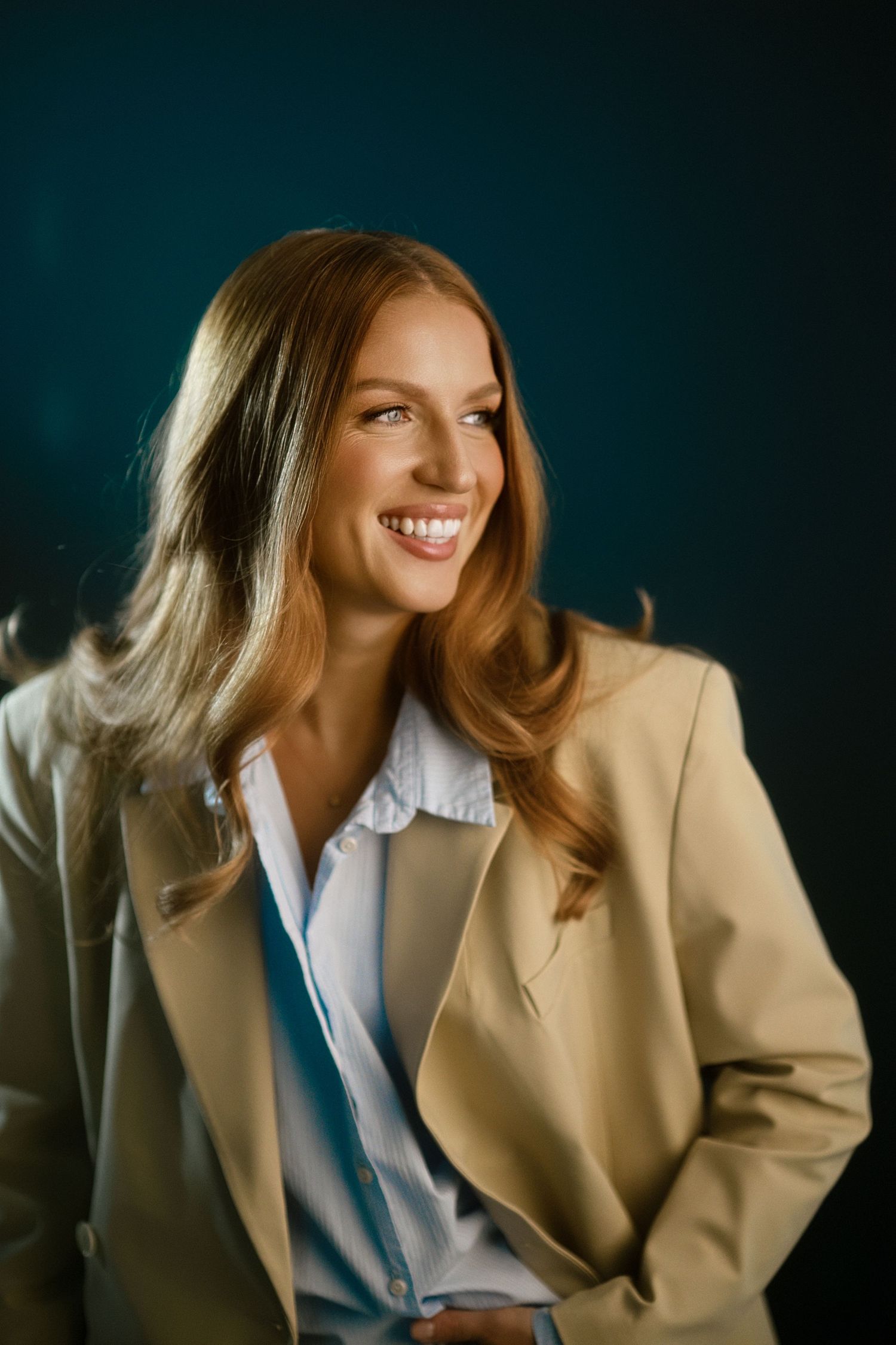 Professional portrait of a person wearing a beige blazer and white shirt, smiling warmly against a dark teal background.