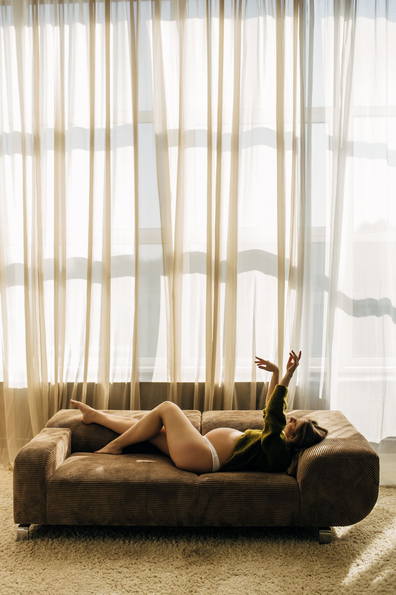 Woman in white lingerie and green top reclining on a brown sofa near sheer curtains in soft natural light.