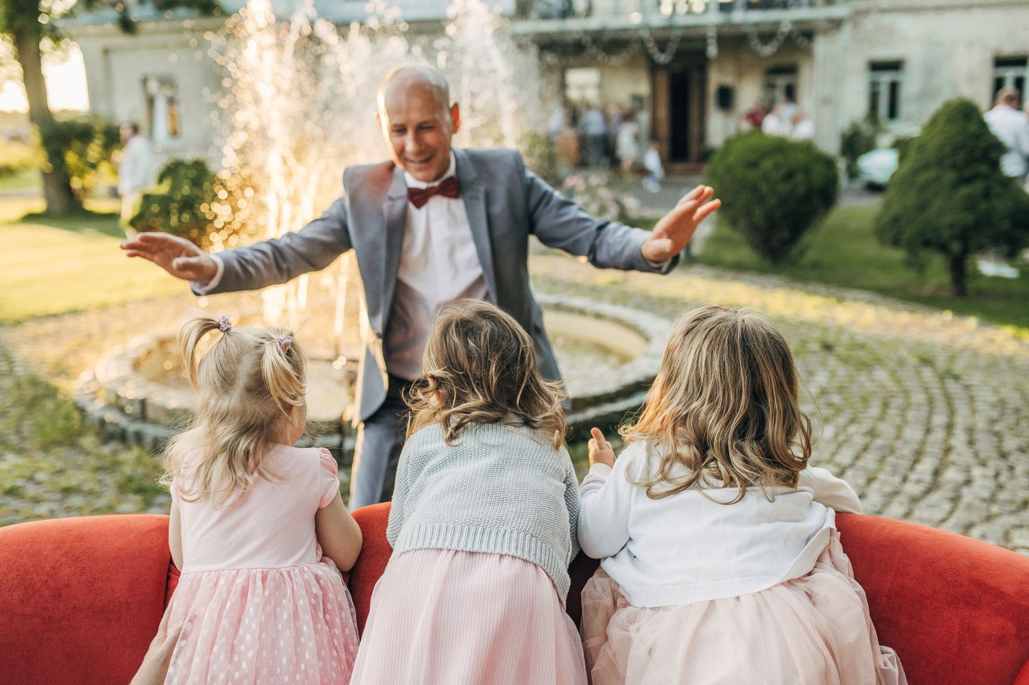 Elderly man in suit joyfully greeting three young girls in pink dresses near a garden fountain.