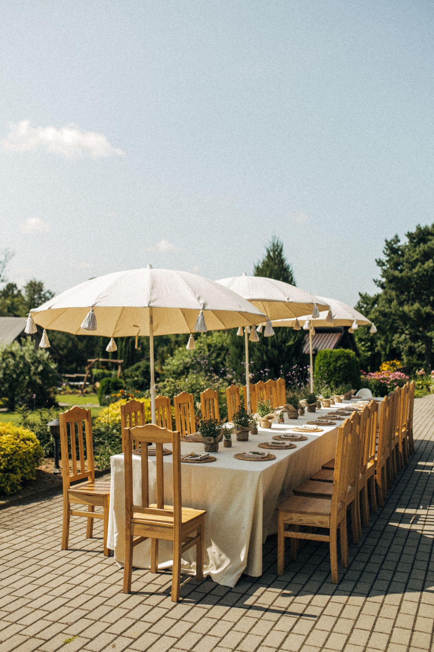 Elegant outdoor wedding reception with long table, wooden chairs, and white parasols in a garden setting.