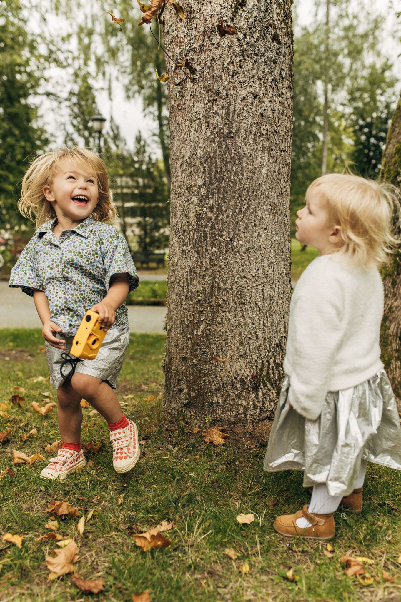 Two toddlers laughing and playing with toy camera near tree in autumn park.