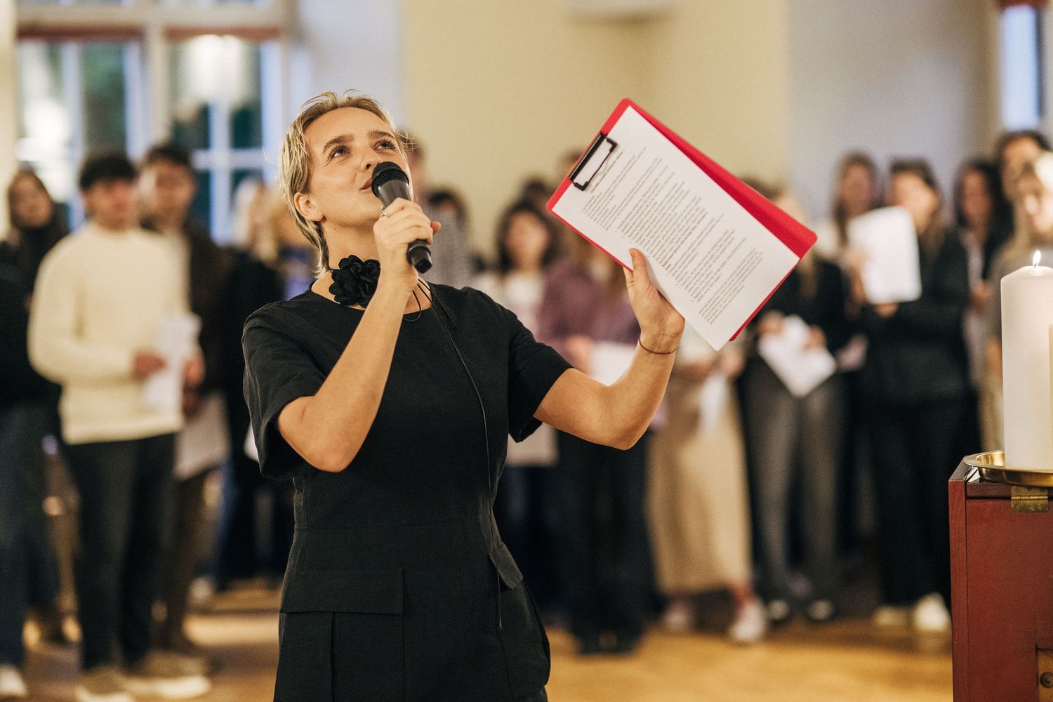 A person in black attire performs at an event with a microphone while an audience watches in the background.