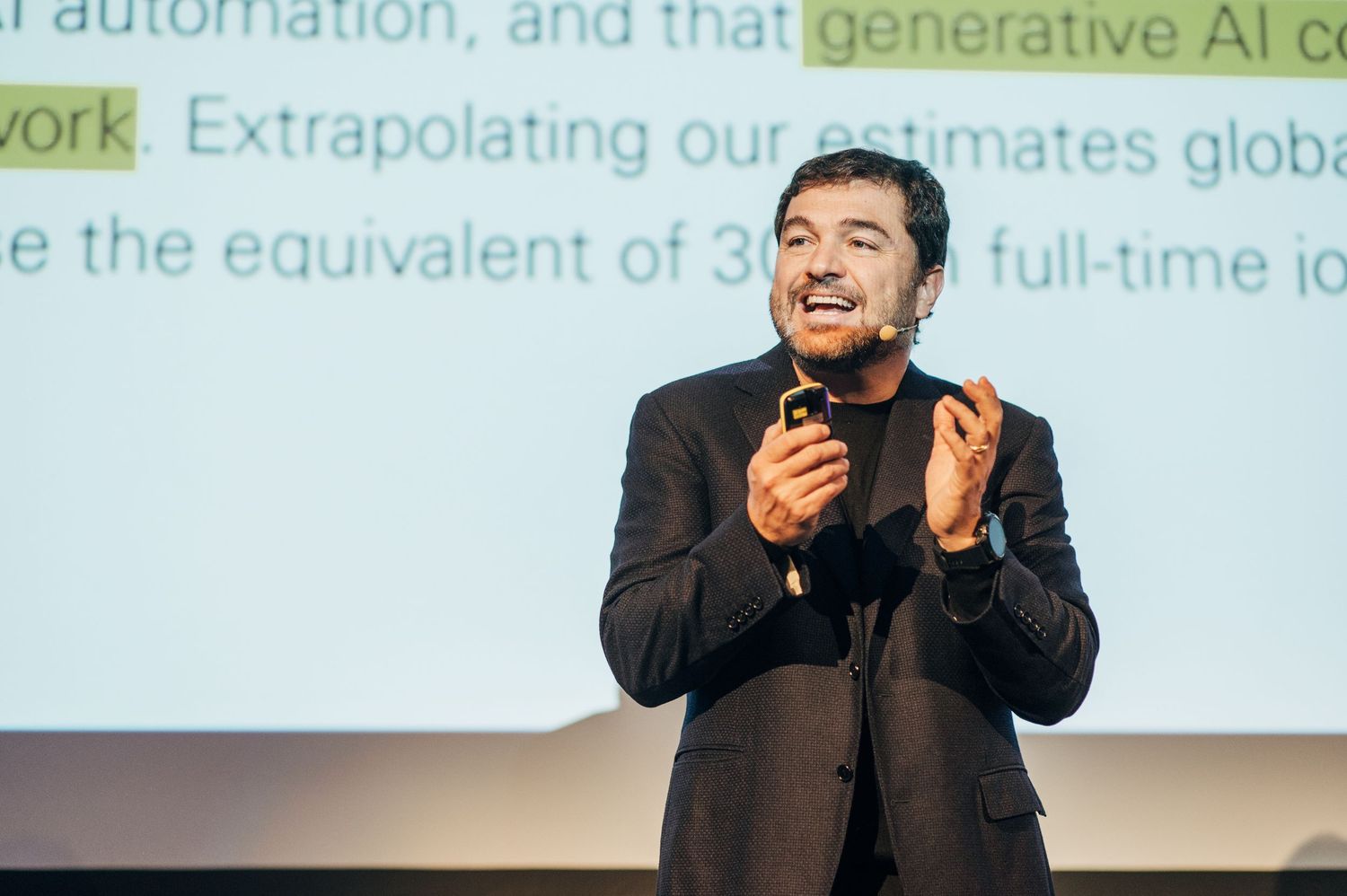 Conference presenter gesturing while speaking in front of projection screen with text.