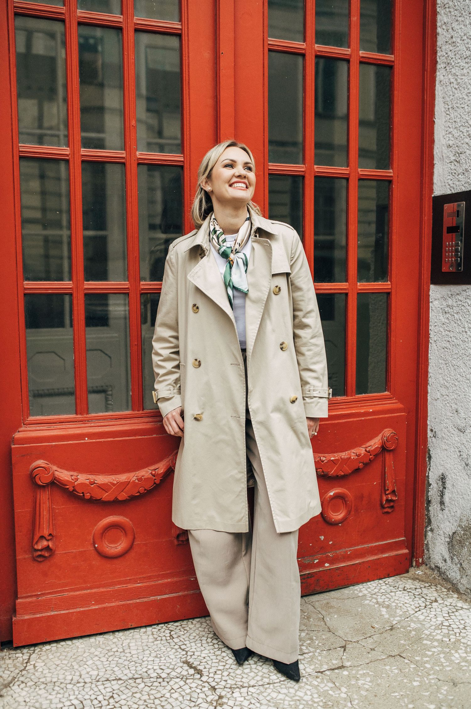 Fashion portrait in casual beige trench coat posing playfully against vibrant red vintage doors in urban setting.