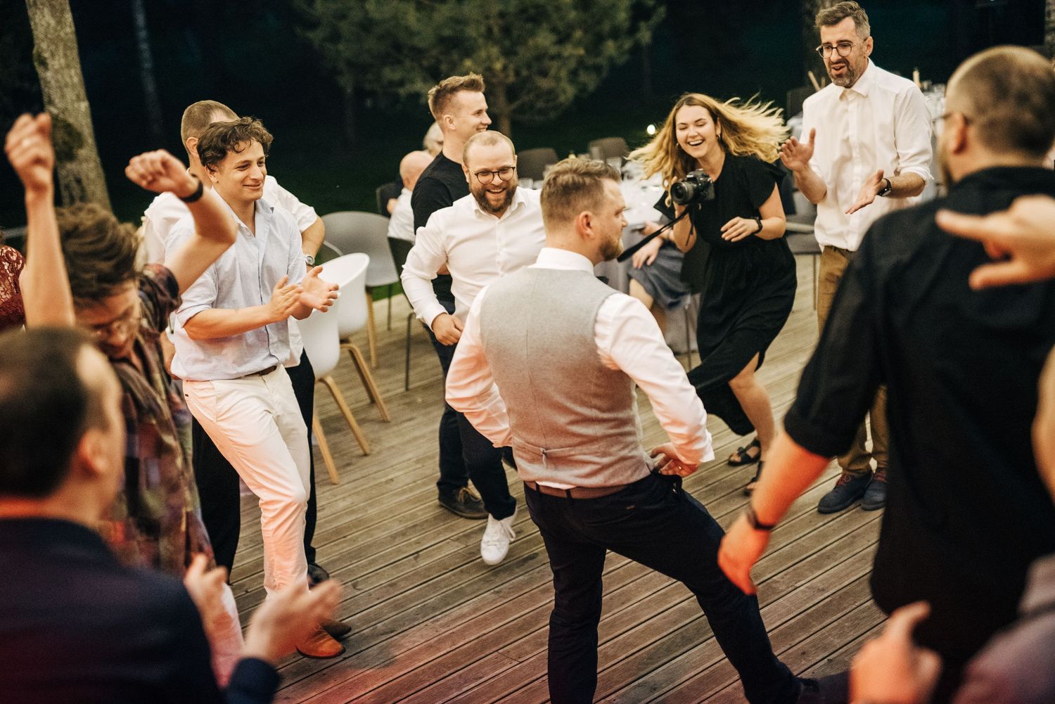 Wedding guests dance energetically on wooden deck during evening reception.