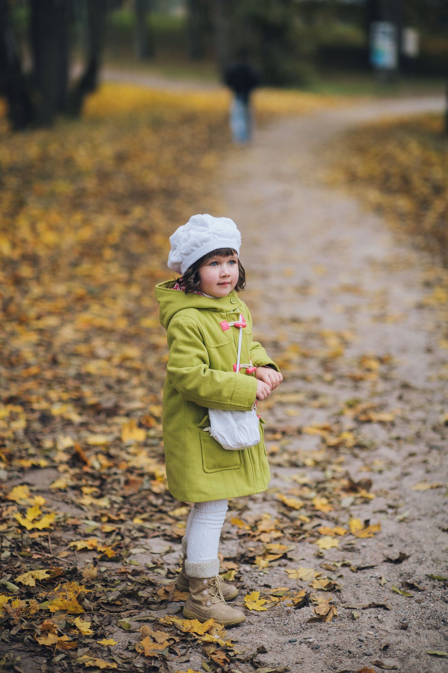 A young girl in a green coat and white beret stands on a leaf-covered autumn park path.