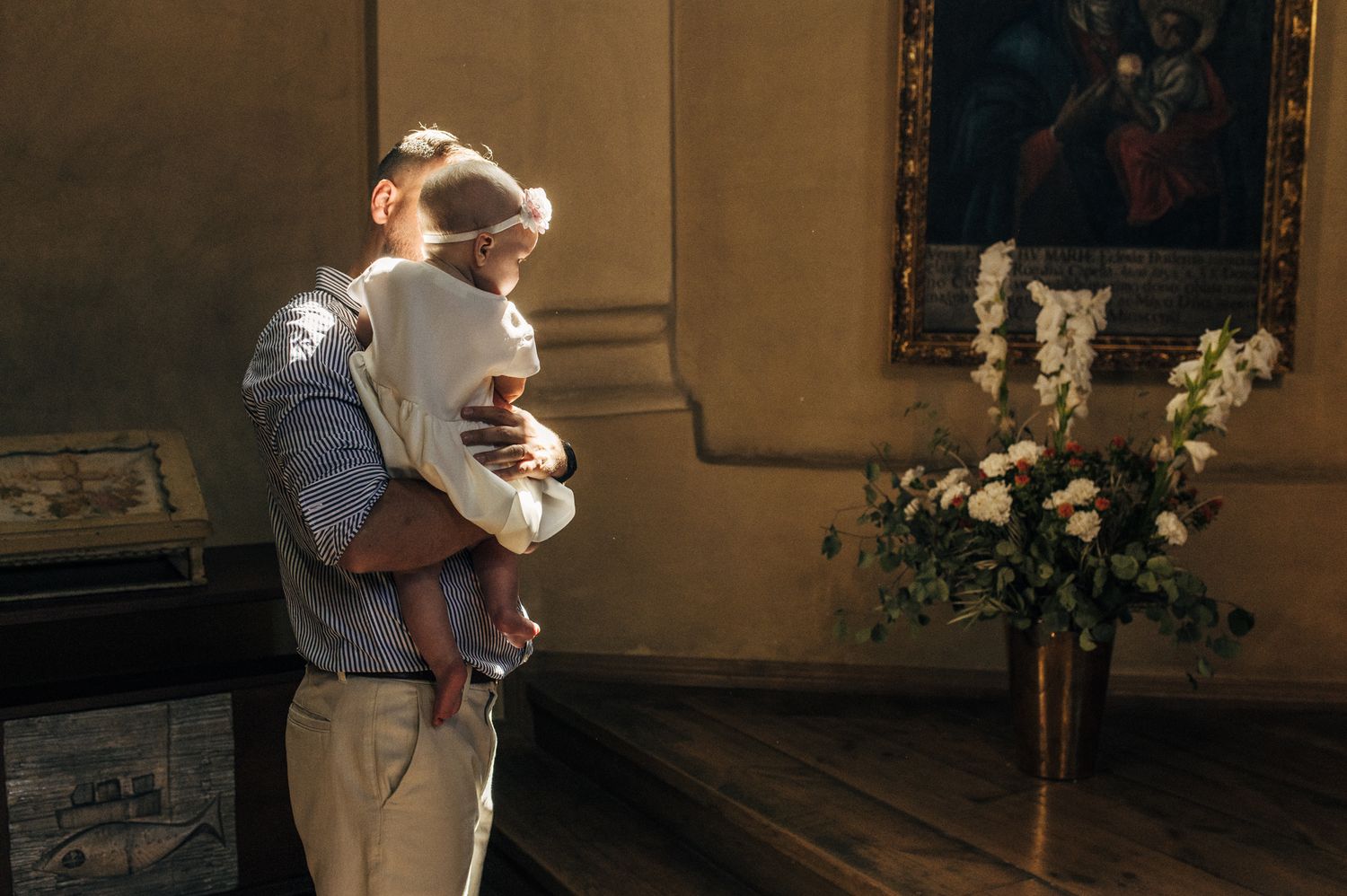 A man tenderly holds a baby wrapped in white during a church baptism ceremony with flowers nearby.