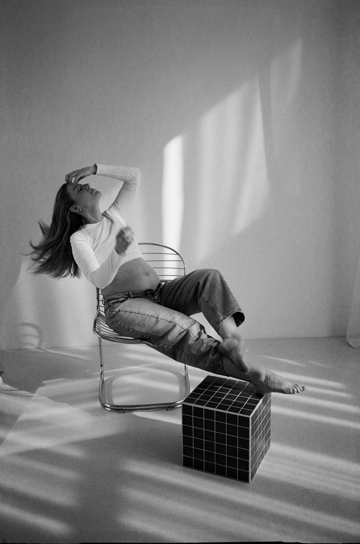 Black and white photo of a woman relaxing in a modern chair, wearing jeans and a crop top, with a geometric cube beside her.