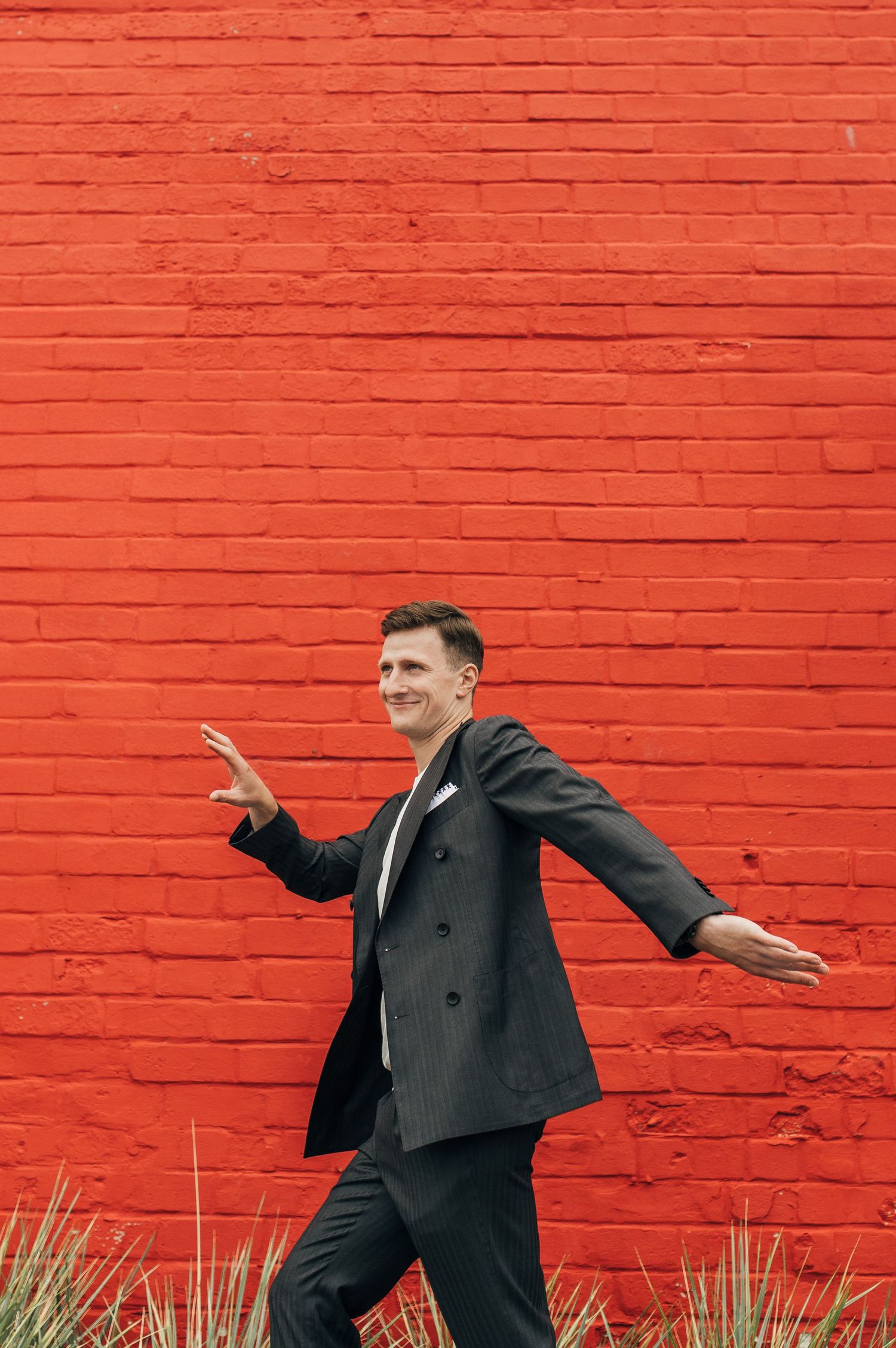 Groom poses playfully in front of vibrant red brick wall.