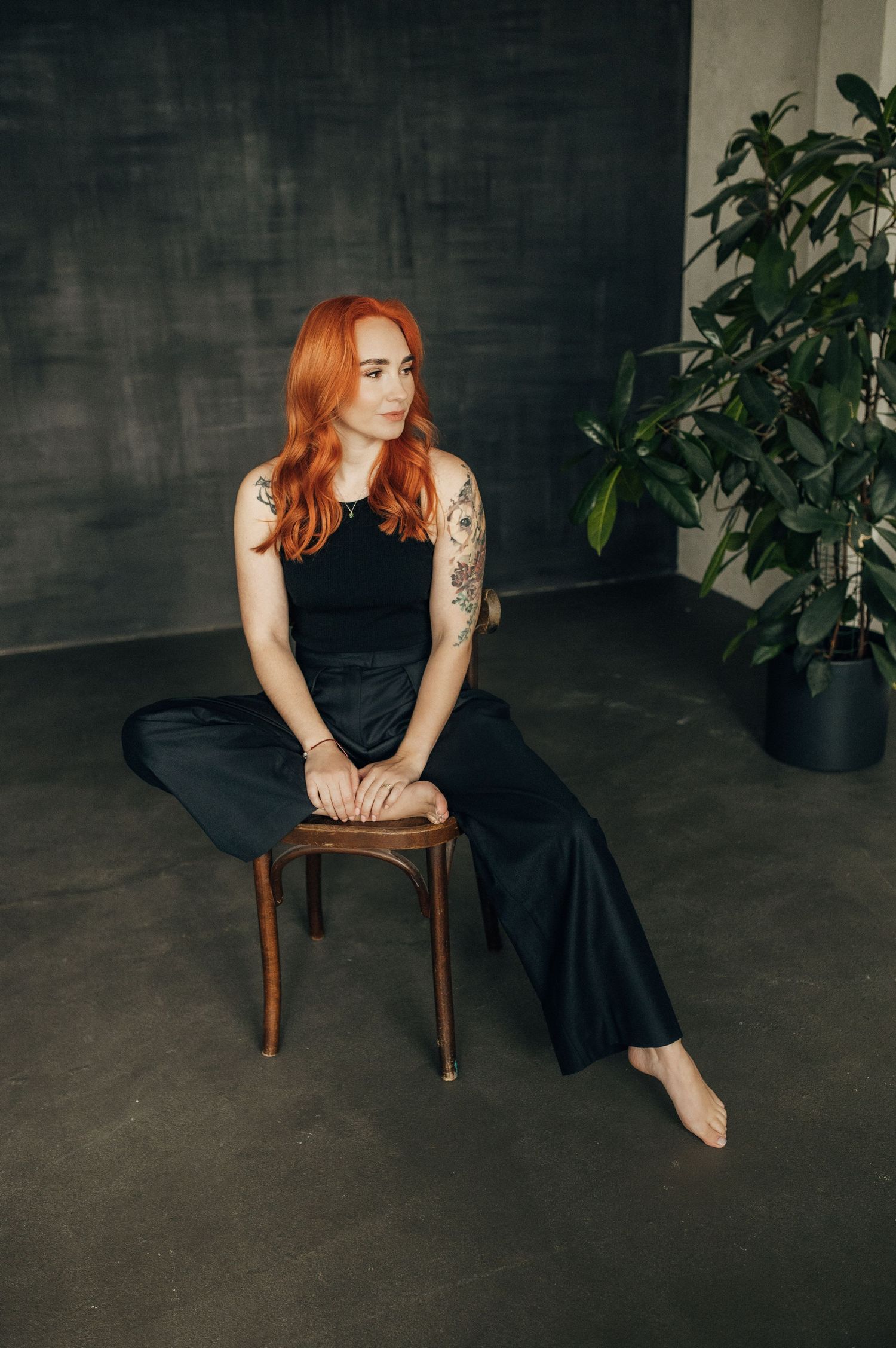 Moody studio portraits with wooden chair, plants and dark background featuring black dress.