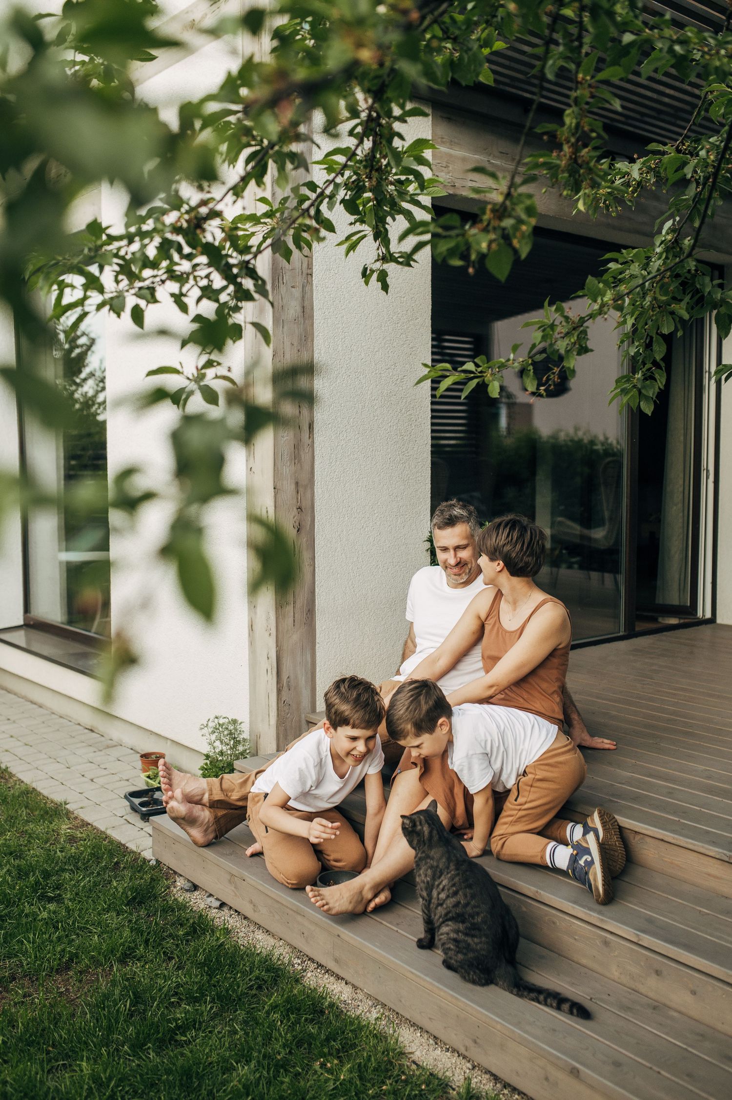 Family relaxing together on wooden deck porch with green foliage in background.