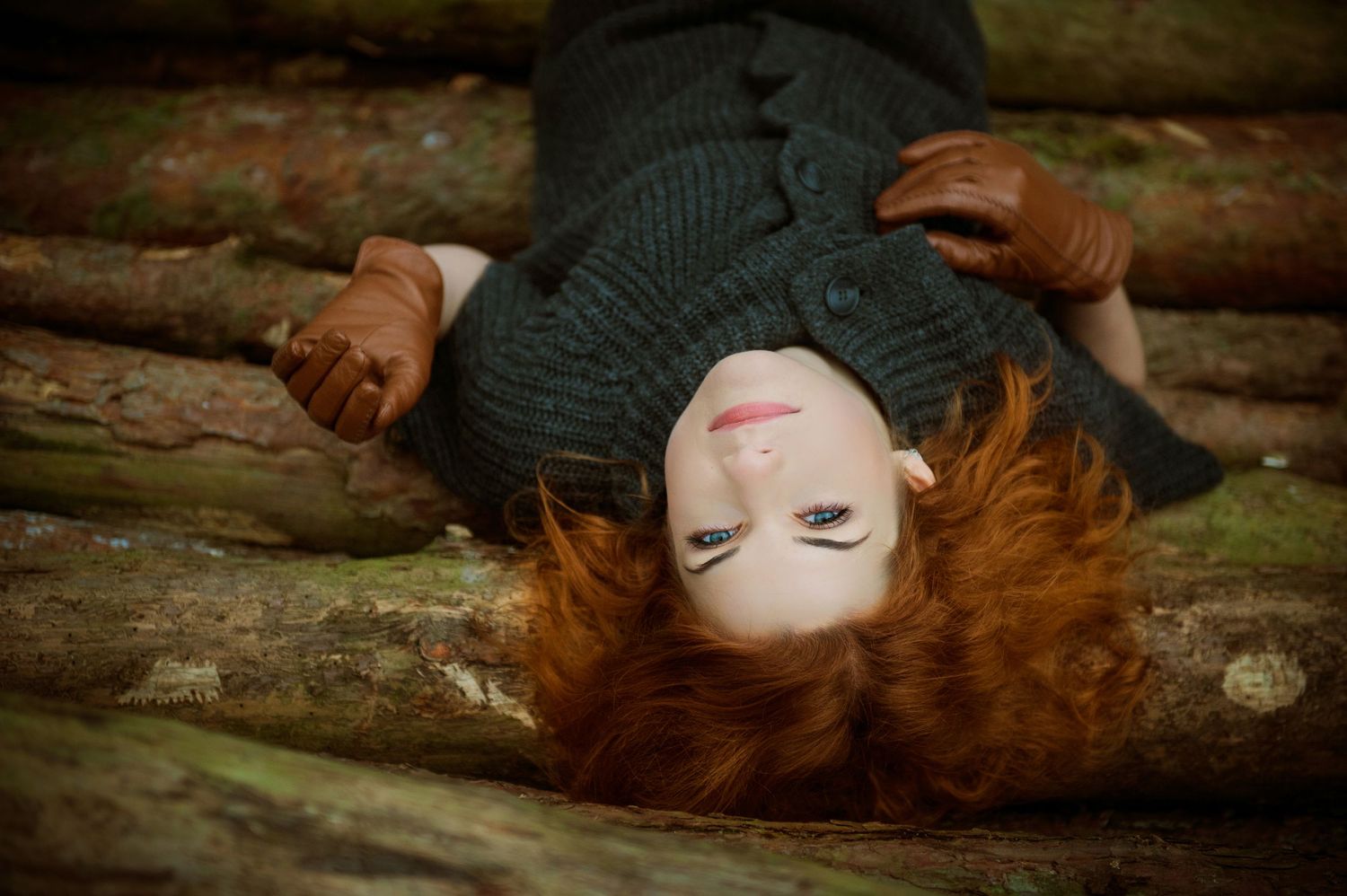Person with red hair lying on wooden logs in dark winter clothing.