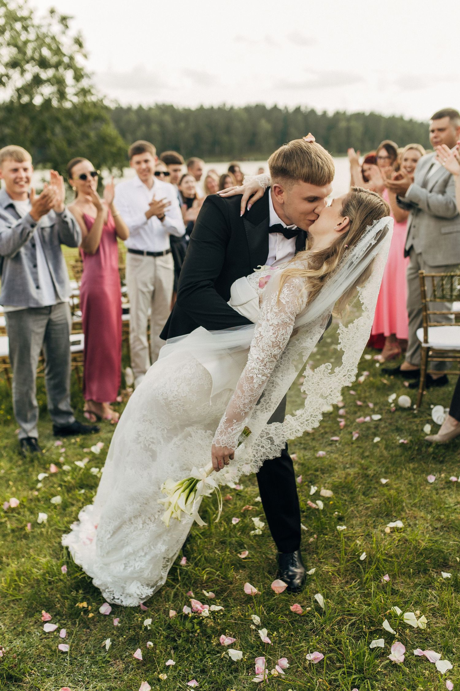 Couple performs dramatic dip dance move surrounded by wedding guests on grassy field with scattered flower petals.