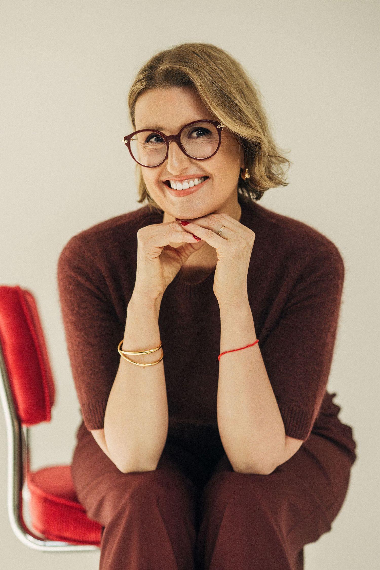 Smiling person in burgundy sweater and glasses rests chin on hands while sitting in front of red chair.