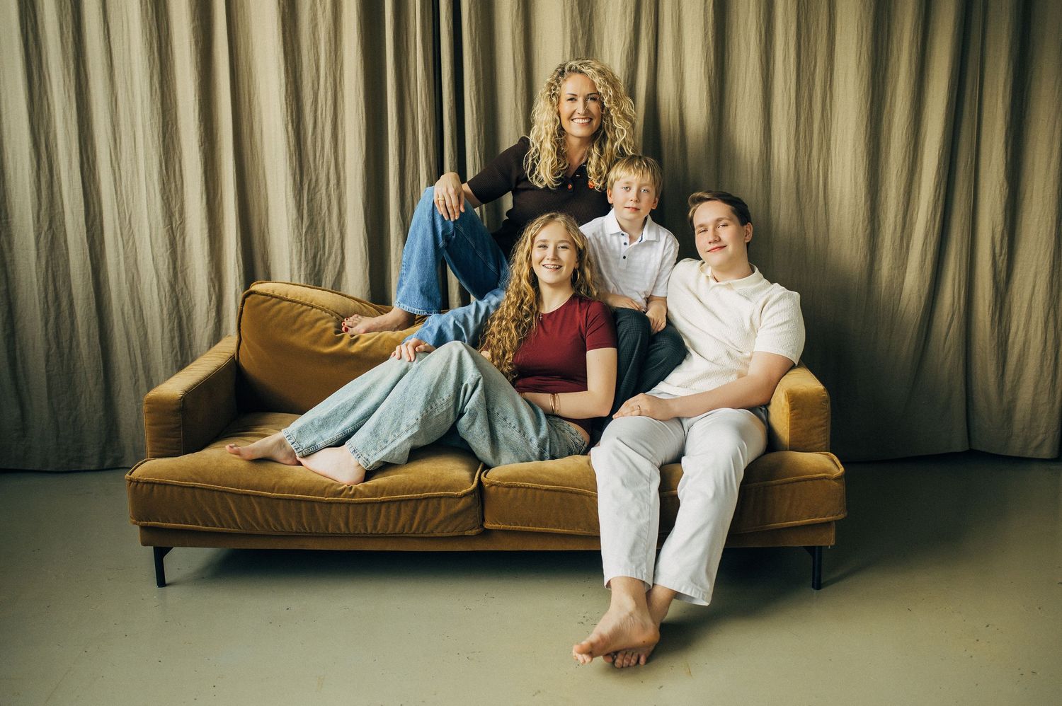 Family of four posing together on a golden velvet sofa against a neutral curtain backdrop in a professional photo shoot.