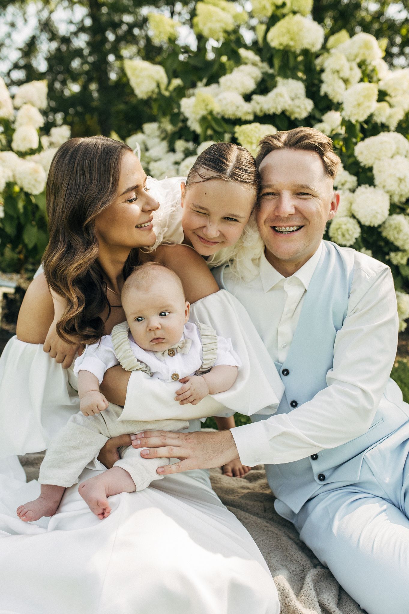 Happy family of four posing outdoors with white hydrangeas, parents holding baby and young girl smiling.
