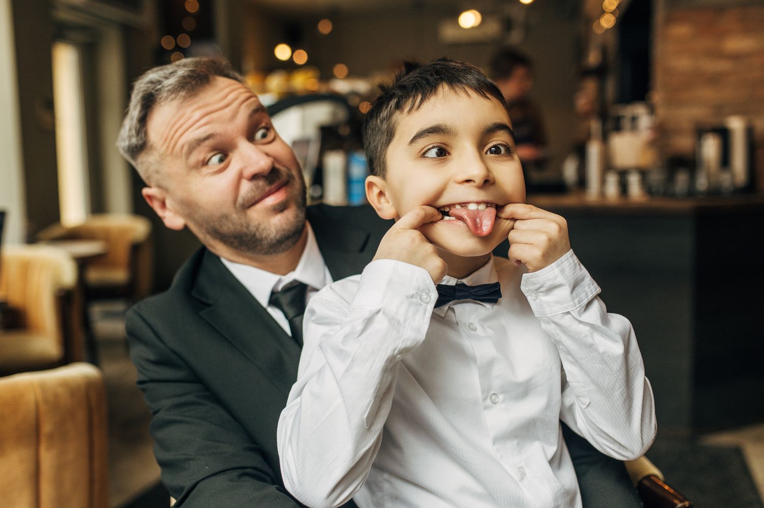 A father and young son make silly faces together, with the boy pulling his cheeks and sticking out his tongue.