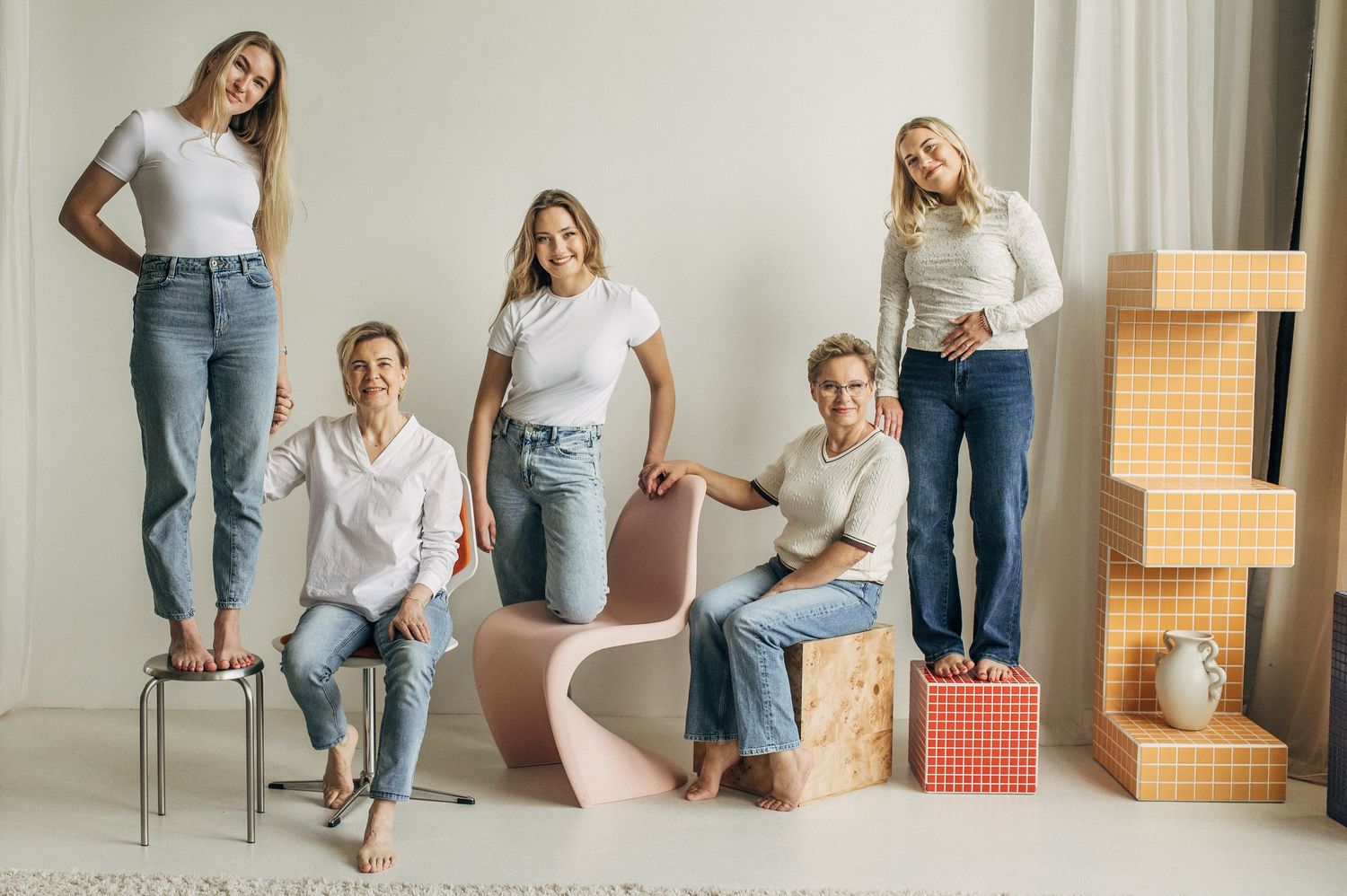 Five women in white tops and jeans pose together in a bright studio with pastel furniture and stacked boxes.