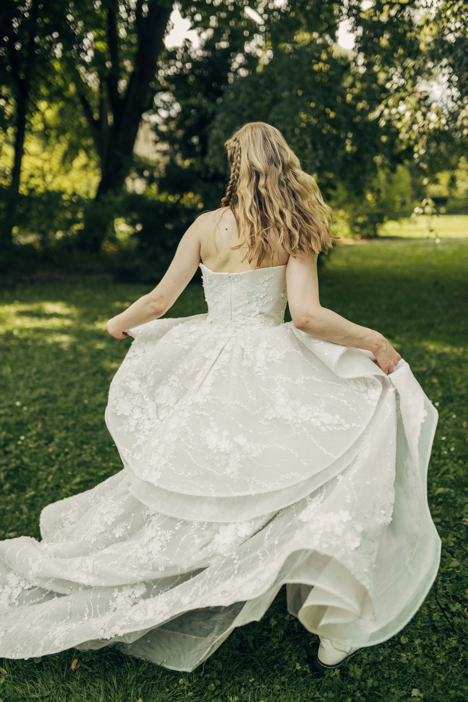 Bride in a flowing white lace wedding gown walks through a lush green garden, holding her dress train.